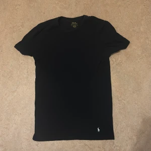 Svart t-shirt från Polo Ralph Lauren - En stilren svart t-shirt från Polo Ralph Lauren med klassisk passform och korta ärmar. T-shirten har en liten vit broderad logga nertill på framsidan. Tillverkad i mjuk bomull som känns skön mot huden. Perfekt till jeans eller shorts.