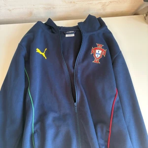 Portugal hoodie jacka från Puma - Mörkblå hoodie jacka från Puma med Portugals landslagsemblem på bröstet och gul Puma-logga. Dragkedja framtill, huva och färgade detaljer i grönt och rött längs sidorna. Stor gul text 'PORTUGAL' på ryggen. Perfekt för fotbollsfans!