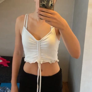 Vit croppad linnetopp - Superfin vit croppad linnetopp från FB Sister med dubbla smala axelband och justerbar snörning framtill. Toppen har v-ringning och volangkant nertill för en söt touch. Perfekt för varma dagar och enkel att styla med jeans eller kjol.