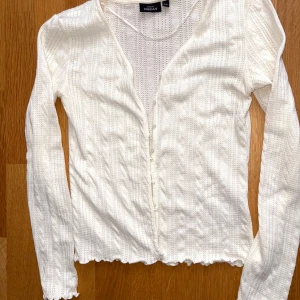 Vit kofta med knappar från KappAhl - Supersöt vit kofta från KappAhl i stickad bomull med diskret hålmönster och smala ränder. Långa ärmar, v-ringad och knappar framtill. Fina volangdetaljer vid ärmslut och nederkant ger en extra touch. Perfekt att slänga över en topp eller klänning.