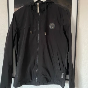 Svart vindjacka från Philipp Plein (S) - Snygg original vindjacka från exklusiva Philipp Plein med PP78-loggor fram och bak. Modellen har en sportig och stilren design i helsvart, med luva, hel dragkedja och två fickor. Unikt tryck baktill med tre hexagon-loggor samt “Plein”-text längst ner.  🔹 Storlek: Small (S) 🔹 Material: 100% polyester (skalet & fodret) 🔹 Tillverkad i: Turkiet 🔹 Skick: Begagnad i bra skick – inga hål eller stora defekter. Något slitage på trycket baktill (se bild).  🔹 Nypris: ca 12 000–13 000 kr 