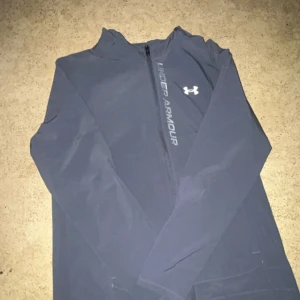 Grå vindjacka från Under Armour  - Snygg grå vindjacka från Under Armour med huva och dragkedja snett över bröstet. Jackan har logga och text på framsidan, meshfoder och långa ärmar. Perfekt för träning eller när du vill ha en sportig look.