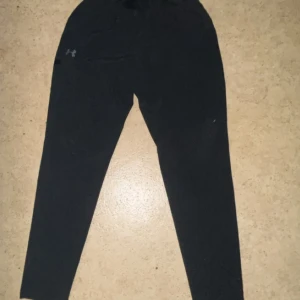 Svarta byxor från Under Armour - Svarta mjukisbyxor från Under Armour med smal passform och elastisk midja med snörning. Byxorna har diskret logga på ena benet och är perfekta för chill eller träning. Tillverkade i mjukt material som känns skönt mot huden.