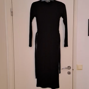 Svart ribbad långklänning NA-KD XS - Svart långklänning från NA-KD Reborn i ribbad stickad kvalitet. Klänningen har lång ärm, rund hals och knytband i midjan för en snygg siluett. Perfekt för dig som gillar en stilren och bekväm look. Nertill på klänningen finns det slits på båda sidor