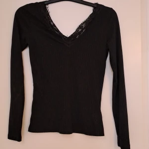 Svart ribbad topp med spets från NA-KD - Svart långärmad topp från NA-KD i ribbat material med snygg v-ringning och spetsdetaljer runt halsen. Figurnära passform som ger en stilren look. Perfekt att styla med jeans eller kjol för en trendig outfit. 