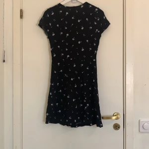 Svart blommig kortklänning H&M🪻 - Supersöt svart kortklänning från H&M med små lila och rosa blommor. Klänningen har korta ärmar, knappar framtill och en lätt utställd passform. Perfekt för dig som gillar en feminin och chill stil.💓