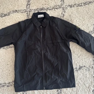 Svart overshirt från Stone Island - Svart overshirt från Stone Island med klassisk krage, dragkedja framtill och ikonisk logga på ärmen. Jackan är tillverkad i lätt polyester och har en clean, minimalistisk look som passar perfekt till streetwear. Snygg och enkel att matcha med det mesta. För mer info som måttet kan du skicka ett PM. 