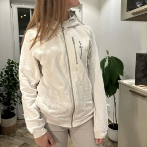 Vit hoodiejacka från Sail Racing - Vit hoodiejacka från Sail Racing med dragkedja framtill och bröstficka med zip. Broderad logga på bröstet och tryck på huvan. Mjuk insida och ribbade muddar. Perfekt för dig som gillar en clean och sportig stil.
