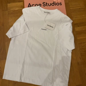 Vit t-shirt från Acne Studios - Clean och stilren vit t-shirt från Acne Studios i 100% bomull. Boxy passform med korta ärmar och rund halsringning. Diskret Acne Studios-logga på bröstet. Perfekt basplagg för en enkel och snygg look.