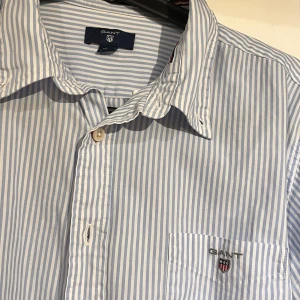 GANT skjorta blå - Snygg skjorta från Gant i blått och vitt med klassiska vertikala ränder. Skjortan har lång ärm, bröstficka med broderad Gant-logga och knäppning framtill. Perfekt för dig som gillar en stilren och fräsch look. Storlek (xs/s)