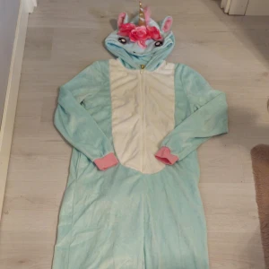 Unicorn onepiece med horn och blommor - Supermysig mintgrönsblå unicorn onepiece med huva, guldigt horn, rosa blommor och öron. Dragkedja framtill, vita detaljer på magen, rosa muddar och fluffig rosa svans bak. Perfekt för dig som älskar enhörningar och vill sticka ut!