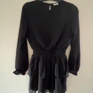 Svart volangklänning från Kappahl - Supersnygg svart kortklänning från Kappahl med långa ärmar och volangkjol. Klänningen har markerad midja och lätt transparenta ärmar med resår vid handlederna. Perfekt för dig som gillar en stilren look med extra volym i kjolen.