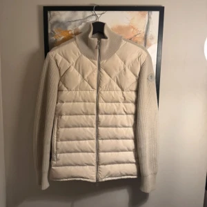  pufferjacka från Moncler - Storlek L men sitter mer som L.Snygg beige pufferjacka från Moncler med quiltad framsida och stickade ärmar och rygg. Jackan har hög krage, dragkedja framtill och Moncler-logga på ärmen. Perfekt för dig som vill ha en stilren och exklusiv look under höst och vinter.