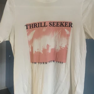 Vit t-shirt med bandtryck Thrill Seeker - Snygg vit t-shirt med tryck i rosa och svart framtill. Motivet föreställer en konsertscen med texten 'THRILL SEEKER' och 'NEW YORK TOUR 1980'. Klassisk passform och rund hals, perfekt för dig som gillar bandtröjor och retrostil.