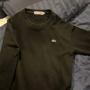 Svart sweatshirt från Lacoste - Svart sweatshirt från Lacoste med klassisk rund halsringning och broderad krokodil-logga på bröstet. Tröjan är i mjuk bomullsmix och har långa ärmar den är också riktigt skön på insidan. Perfekt för en clean och stilren look. Den är L men sitter som M, vid frågor om pris eller annat är det bara att fråga 🙋🏽‍♂️ 😄