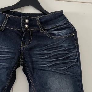 Mörkblå skinny jeans Miss Anna - Snygga mörkblå skinny jeans från Miss Anna med coola skrynkliga detaljer framtill och bak. Jeansen har dubbla knappar i midjan, klassiska fickor och kontrastsömmar. Perfekta för dig som gillar en tajt passform och vill sticka ut lite extra.