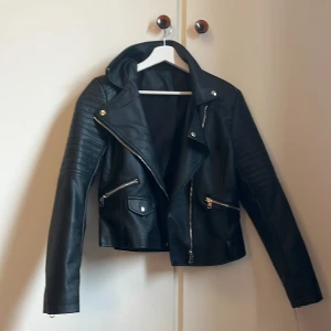 Svart skinnjacka med dragkedjor - Snygg svart skinnjacka med coola detaljer som silvriga dragkedjor och quiltade partier på axlarna. Jackan har en klassisk bikerstil med bred krage och flera fickor framtill. Perfekt för dig som vill ha en edgy look.