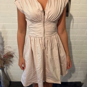 Ljusrosa kortklänning med dragkedja - Supersöt ljusrosa kortklänning med markerad midja och cool dragkedja framtill. Klänningen har v-ringning, ärmlös design och lätt glansigt material som ger en fräsch look. Perfekt för dig som gillar en feminin men edgy stil.