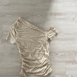 Beige oneshoulder topp från Gina Tricot - Säljer en beige oneshoulder topp från Gina Tricot. Toppen har kort ärm på ena sidan. Köpt men aldrig använd så har bara legat i garderoben. Storlek S🤍🤎