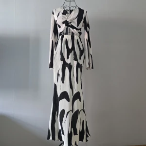 Svartvit långklänning med twistad front - Unik långklänning i svartvitt abstrakt mönster med twistad omlottfront och långa ärmar. Klänningen har en figurnära passform upptill och faller snyggt längs kroppen. Materialet är plisserat och ger en cool struktur. Perfekt för dig som vill sticka ut med en modern look.