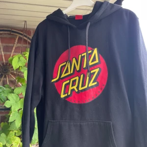 Svart Santa Cruz hoodie med tryck - Svart hoodie från Santa Cruz med stort rött och gult logotryck på bröstet. Klassisk modell med huva, snörning och känguruficka. Perfekt för dig som gillar streetwear och skatekänsla.