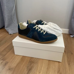 Maison Margiela GAT Sneaker - Snygga sneakers från Maison Margiela i en djupblå färg med detaljer i mocka och skinn. Skorna har vita snören, beige innersida och klassisk gummisula. Perfekt för dig som gillar stilrena och trendiga sneakers med en lyxig känsla. De är helt nya med tillhörande box, dustbags och kvitto. Priset kan diskuteras vid snabb affär🤝