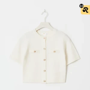 Vit kofta med guldknappar - Kortärmad vit kofta med snygga guldknappar framtill och dekorativa fickor på bröstet. Ribbstickade kanter vid hals, ärmslut och nederkant ger en clean look. Perfekt för lager-på-lager och passar till allt.