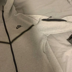 Grå Nike tech tracksuit (Nytt skick) - Snygg grå hoodie från Nike med hel dragkedja och svarta detaljer längs bröstet och ärmen. Jackan har huva, fickor med dragkedja och klassisk Nike-logga på bröstet. Perfekt för chill eller träning, riktigt clean och sportig look.