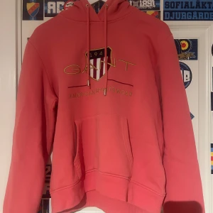 Rosa hoodie från Gant med tryck - Säljer en rosa hoodie från Gant med broderat logotyp-tryck och texten 'American Sportswear' på bröstet. Tröjan har huva med snörning och en stor magficka. Helt ny aldrig använd 