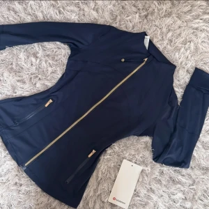Marinblå träningsjacka från Lululemon - Snygg marinblå träningsjacka från Lululemon med figurnära passform, guldfärgade dragkedjor och hög krage. Jackan har långa ärmar och två praktiska fickor med dragkedja framtill. Perfekt för dig som vill ha en sportig och stilren look.