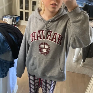 Grå Harvard hoodie med tryck - Säljer en grå hoodie från Harvard med vinrött och vitt tryck framtill. Tröjan har huva med snörning och en stor magficka. Oversized passform och mjukt material, perfekt för chill dagar.