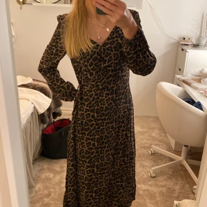 Leopardmönstrad långklänning med knappar - Säljer en långklänning med leopardmönster i bruna och svarta toner. Klänningen har v-ringning, långa ärmar och knappar framtill. Omlottmodell med knyt i midjan, på bild 3 ser man två ringar och det menas med att man kan knäppa den så att den blir två olika storlekar, de gör de att klänningen passar både XS och M/L