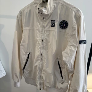 Vit vindjacka från Gant - Snygg vit  vindjacka från Gant med dragkedja framtill och två fickor med svarta detaljer. Jackan har broderade patches på bröstet och ärmen, samt ribbade muddar vid ärmslut och nederkant. Perfekt för dig som gillar klassisk och sportig stil. Storlek: 2XL barn. Passar vuxen S