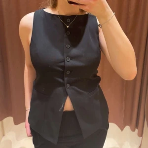 Svart väst med knappar - Snygg svart väst med rak passform och knäppning framtill. Västen har en stilren design med rund halsringning och fem knappar i mitten. 