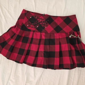 Röd och svart rutig kjol med kedja - Supercool röd och svart rutig kortkjol från VistWeens med plisserad design, dragkedja bak och silverkedja som detalj. Kjolen har tryck och små nitar framtill för extra edge. Perfekt för dig som gillar alternativ stil!