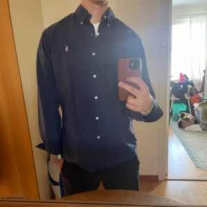 Mörkblå skjorta från Ralph Lauren M - Säljer en mörkblå skjorta från Ralph Lauren i slim fit med klassisk button down-krage. Skjortan är i 100% bomull med stretch och har vita knappar samt den ikoniska rosa broderade loggan på bröstet. Perfekt för en clean och stilren look. Passar en som är emellan 170-180 cm lång. Hör av er vid intressen!