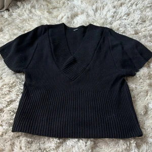 Svart stickad v-ringad tröja - Svart stickad tröja med bred v-ringning och korta ärmar. Tröjan har ribbade detaljer vid halsringning och nederkant, vilket ger en snygg och modern look. Köpt på secondhand! Använd 1 gång.