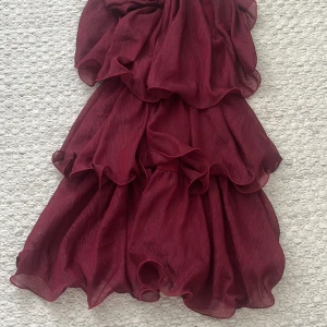 Vinröd volangklänning med axelband - Supersöt vinröd kortklänning med tunna axelband och flera lager volanger. Klänningen har en lätt glansig yta och är gjord i ett tunt, flowy material som ger en riktigt snygg siluett. Perfekt för dig som vill sticka ut med en romantisk vibe.