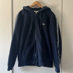 Mörkblå hoodie från Lacoste med huva - Snygg mörkblå hoodie från Lacoste med dragkedja, huva och vita logoband längs ärmarna. Klassisk krokodil-logga på bröstet och stora fickor framtill. Perfekt för dig som gillar sportig och avslappnad stil.