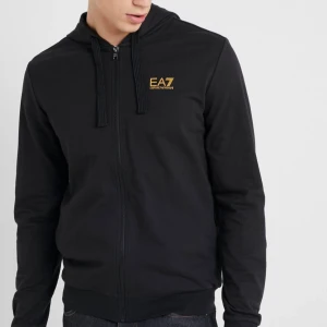 Svart hoodie från Emporio Armani - Säljer en stilren svart hoodie från Emporio Armani med dragkedja och guldfärgad logga på bröstet. Perfekt för en casual look med sin bekväma passform och praktiska huva. Ny pris: 1500kr mit pris: endast 449kr
