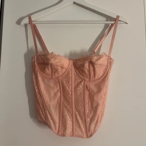 Rosa korsett med spetsdetaljer - Säljer en söt rosa korsett med spetsdetaljer och justerbara axelband. Den har en vacker design med spets över bysten och längs sidorna, 