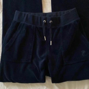 Mörkblå mjukisbyxor från Juicy Couture - Snygga mörkblå mjukisbyxor från Juicy Couture med snörning i midjan och praktiska fickor både fram och bak. Byxorna har en mjuk och bekväm passform, perfekt för en avslappnad stil. Detaljer som knappar på bakfickorna ger en extra touch. Storlek xxs!🥰