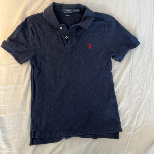 Mörkblå pikétröja från Ralph Lauren - Säljer en snygg mörkblå pikétröja från Ralph Lauren med klassisk krage och knappslå. Tröjan har en röd broderad logotyp på bröstet och är tillverkad i 100% bomull för en bekväm passform. Perfekt för en stilren look! Medela vid minsta funderingar! 👍