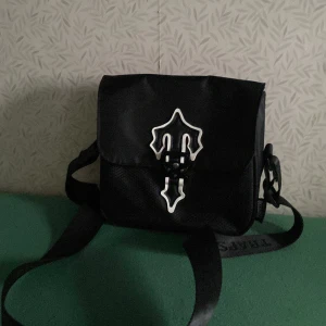 Svart axelväska med dekorativt emblem - Snygg svart axelväska med ett unikt dekorativt emblem på framsidan. Väskan har en justerbar axelrem och ett huvudfack med dragkedja för säker förvaring. Perfekt för att bära dina dagliga nödvändigheter med stil. Passar bra nu när sommaren är nära. Väskan kostar 550kr men man kan pruta i dm om det är en acceptabel pris.