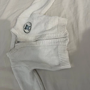 Vit stickad kofta med dragkedja - Säljer en söt vit stickad kofta med dragkedja och ett broderat motiv på bröstet. Koftan är tillverkad i 100% bomull och har ribbade kanter för extra komfort. Perfekt för de kallare dagarna!