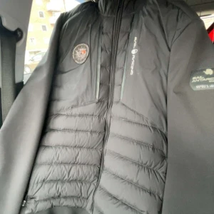 Svart jacka från Sail Racing - Snygg svart jacka från Sail Racing med dragkedja och broderade detaljer. Jackan har en quiltad design på framsidan och märkesloggor på bröstet och ärmen. Perfekt för kyligare dagar.