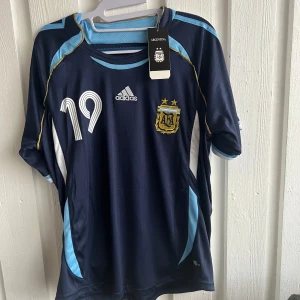Blå Argentina fotbollströja från Adidas - Säljer en blå Argentina fotbollströja från Adidas med nummer 19 och namn på ryggen. Tröjan har korta ärmar och detaljer i ljusblått och vitt. Perfekt för fotbollsfans!