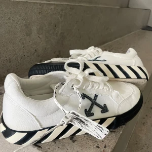 Vita Off-White Sneakers - Snygga vita sneakers från Off-White med svarta och vita ränder på sulan och en orange detalj på sidan. Skorna har snörning och Off-White-logotypen på sidan. Perfekta för en stilren look. Orginal box kommer med!