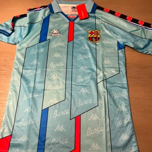 Retro Barcelona fotbollströja från Kappa - Snygg retro Barcelona fotbollströja från Kappa i ljusblått med mönster av klubbens namn och logga. Tröjan har korta ärmar och en klassisk krage med röd och blå detalj. På ryggen finns namnet 'Ronaldo' och nummer 9. Perfekt för samlare eller fans av klassisk fotbollshistoria.
