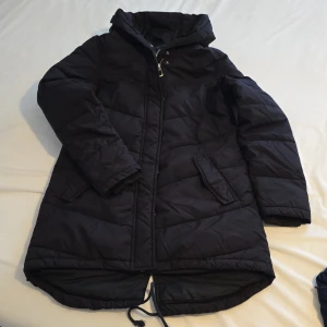 Svart dunjacka från Everest - Säljer en svart dunjacka från Everest med dragkedja och knappar. Jackan har en långärmad design och en praktisk huva. Perfekt för kyliga dagar med sin quiltade stil och bekväma passform.